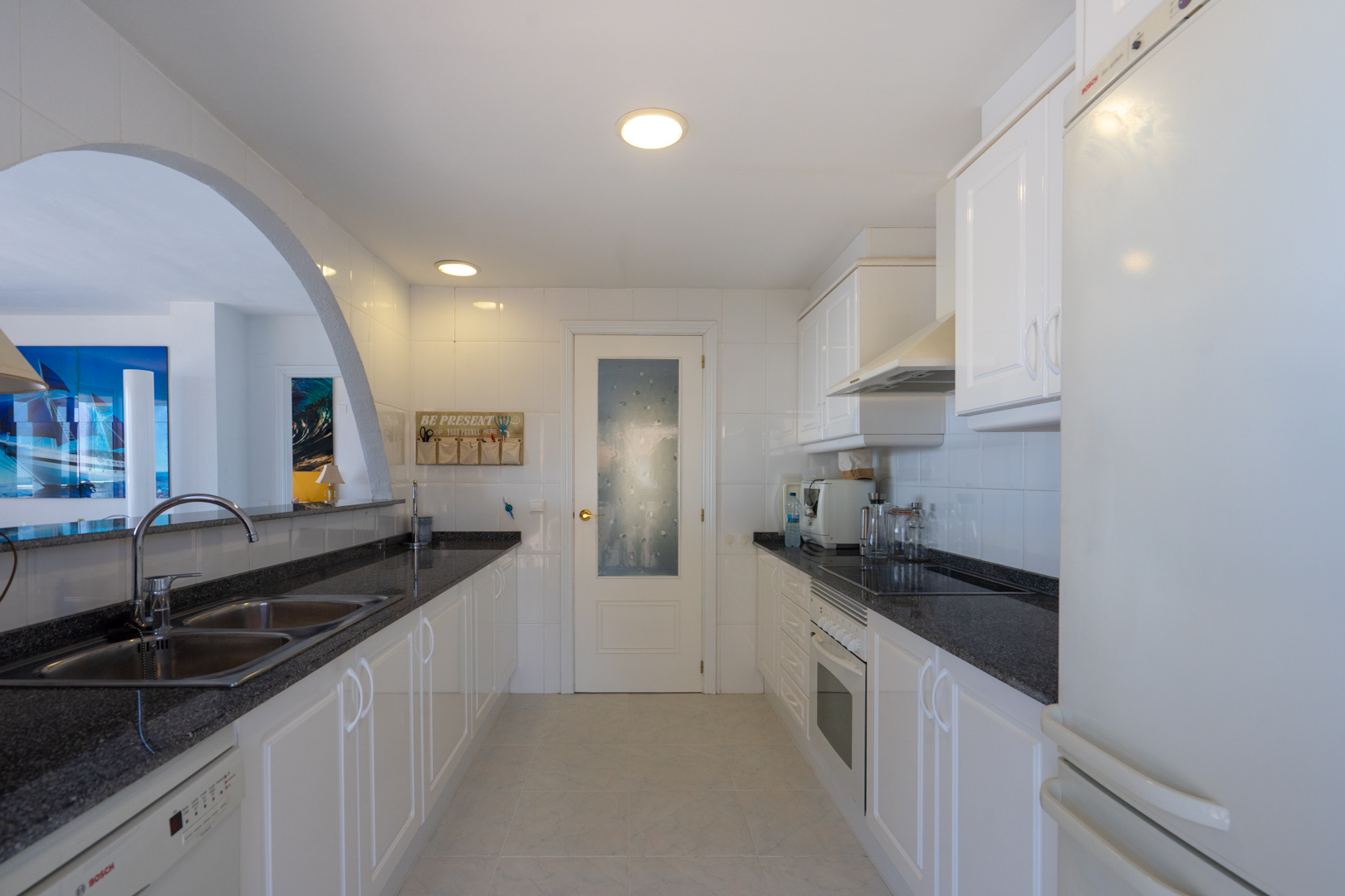 Cocina funcional con mobiliario blanco, encimeras de granito oscuro y electrodomésticos integrados, conectada a la zona de estar mediante un arco.