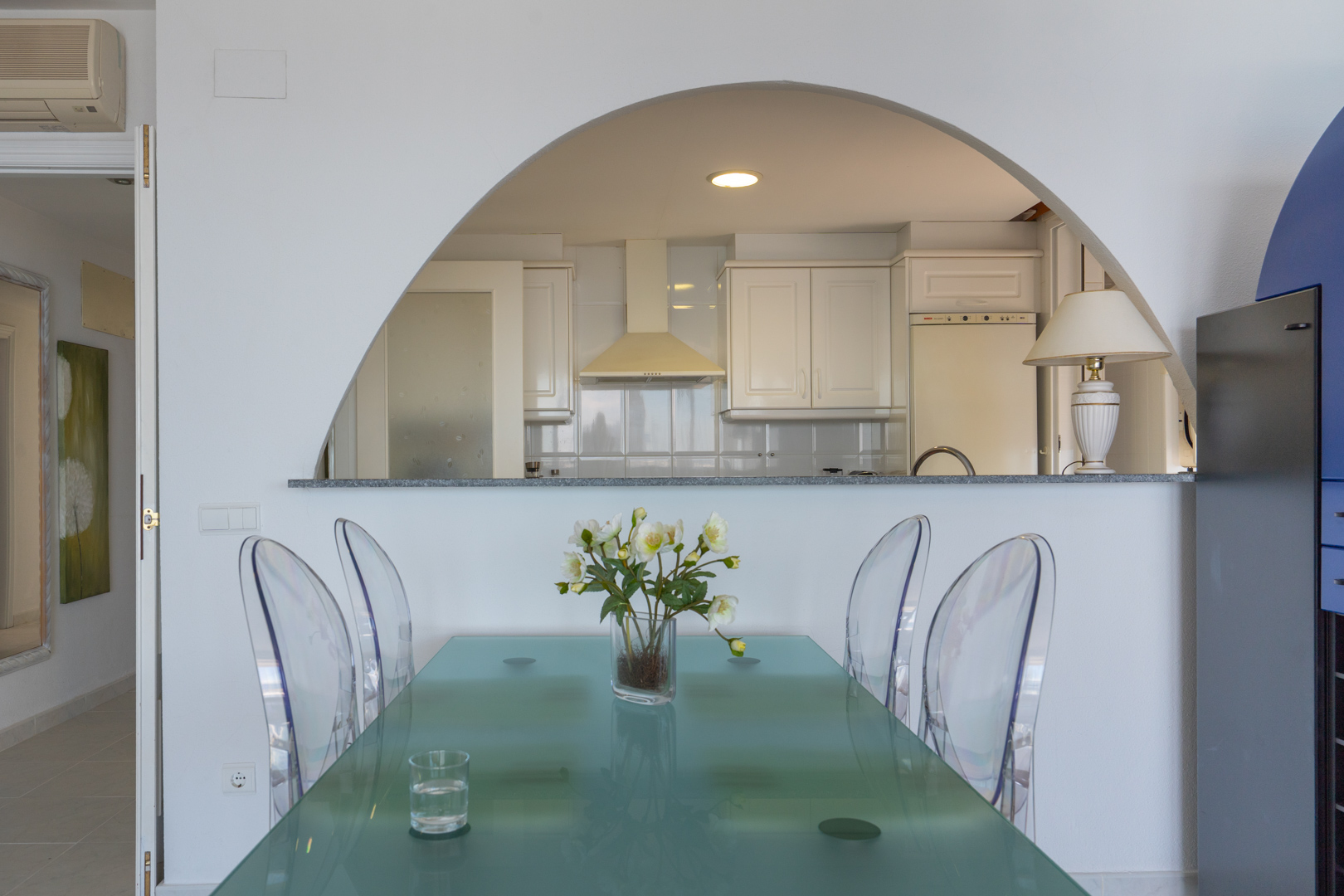 Comedor con mesa de cristal esmerilado y sillas transparentes, conectado a una cocina de armarios blancos mediante un arco y barra de granito gris. Decorado con flores blancas.