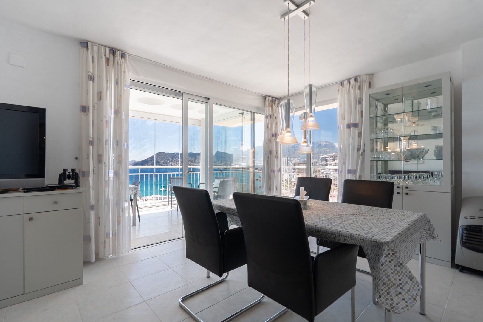Comedor luminoso con mesa moderna y sillas, acceso directo a terraza acristalada con vistas panorámicas al mar y al Peñón de Ifach. Vitrina con cristalería.