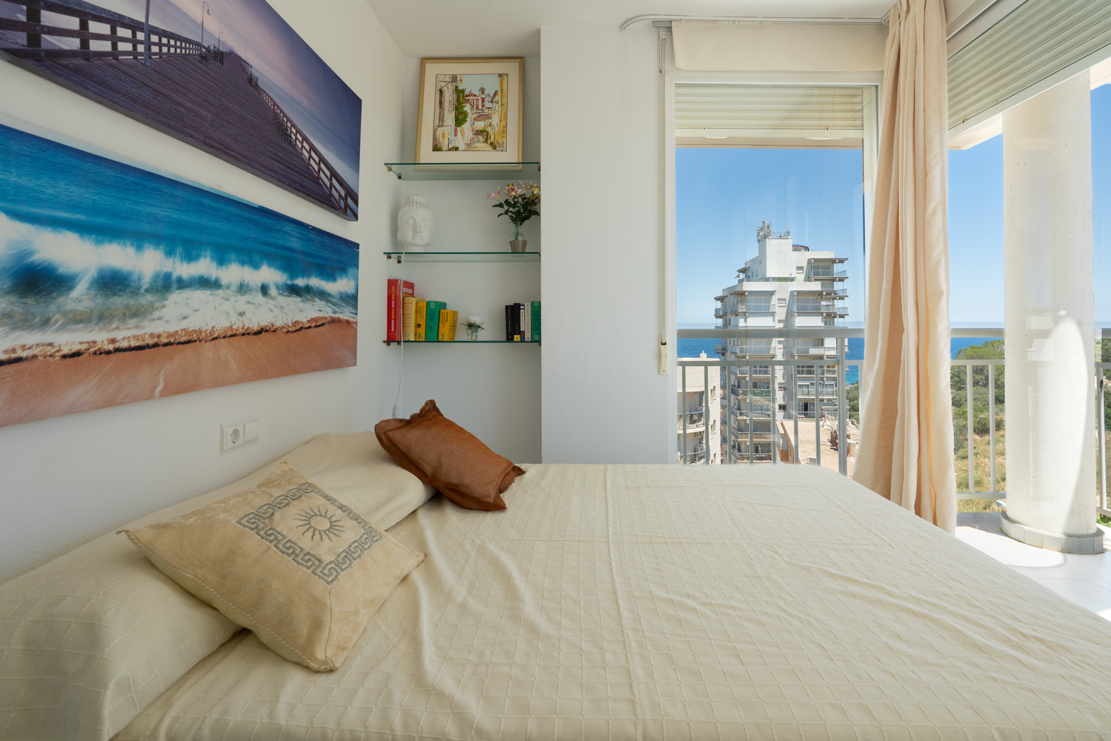 Dormitorio con cama vestida, decorado con láminas marinas y estanterías de cristal. Acceso a balcón con vistas parciales al mar y a un edificio moderno.