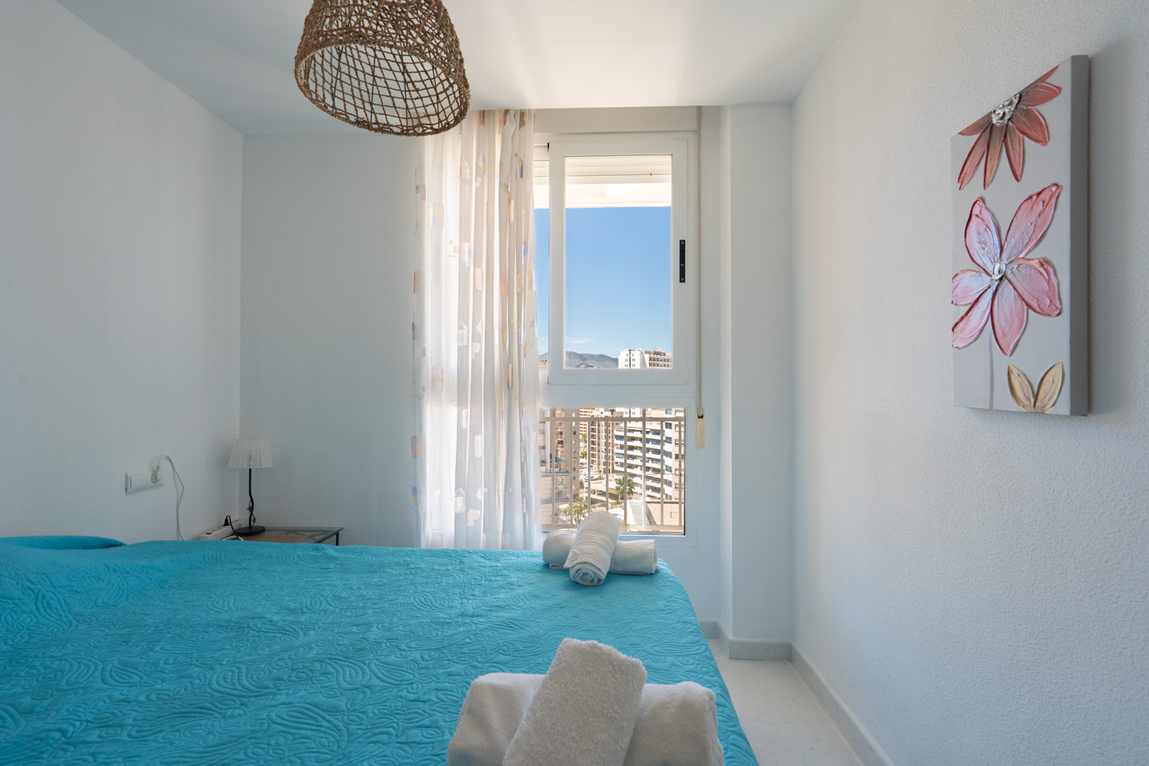 Dormitorio luminoso con cama de edredón azul y toallas blancas. Dispone de una ventana con vistas urbanas y montañosas, lámpara de techo de mimbre y un cuadro floral.