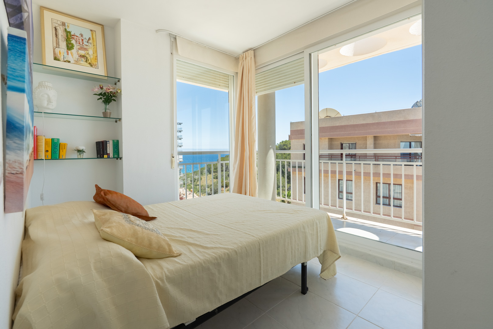 Dormitorio luminoso con cama de matrimonio, estanterías de cristal decoradas y acceso directo a una terraza con vistas parciales al mar y al entorno urbano.
