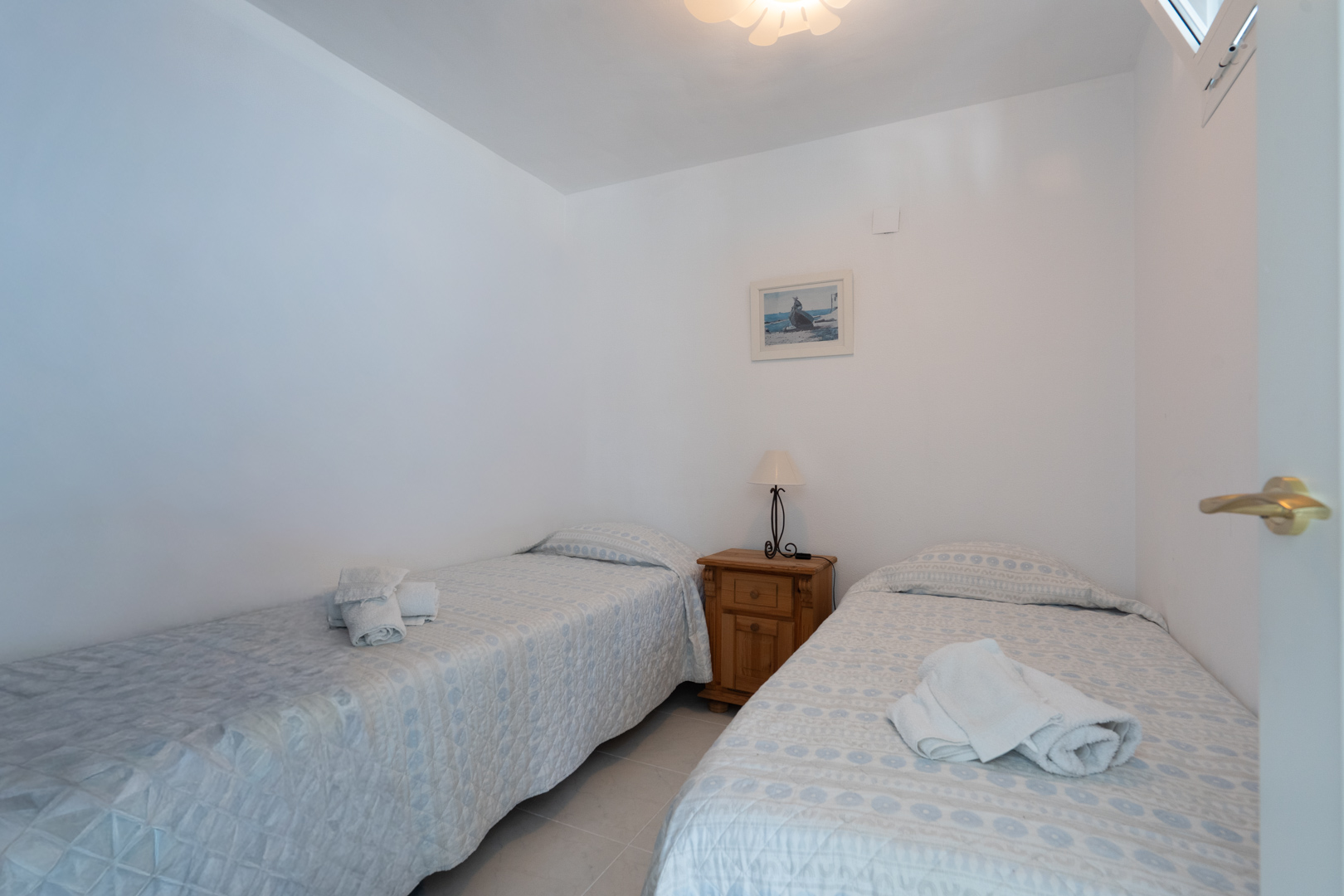 Dormitorio luminoso con dos camas individuales vestidas con colchas claras, una mesita de noche de madera y una lámpara, complementado por un cuadro decorativo y una ventana superior.