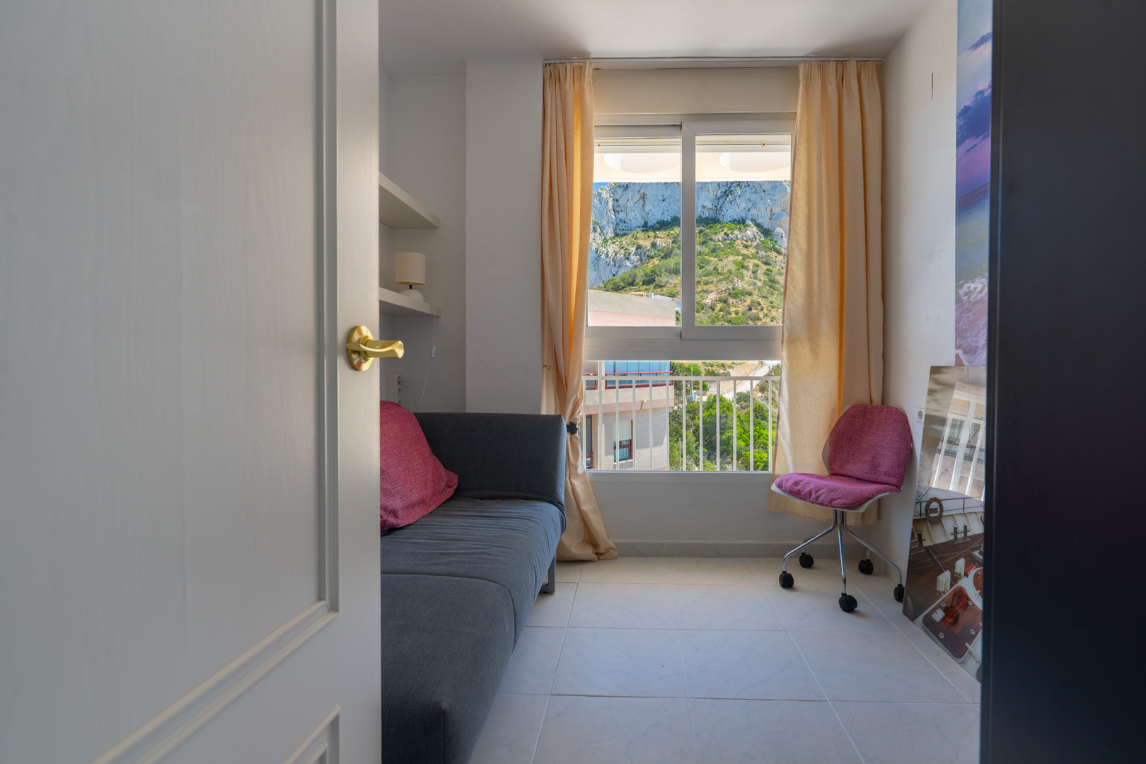 Dormitorio secundario o despacho con sofá y vistas al Peñón de Ifach. Dispone de estanterías integradas y suelo de baldosas claras.