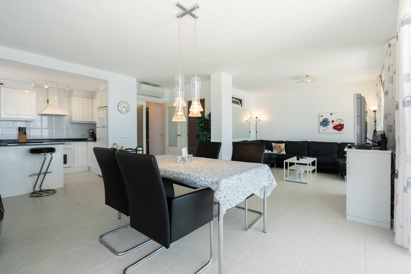 Espacio diáfano con cocina blanca, barra americana, comedor con lámpara colgante de tres luces y salón con sofá seccional negro.