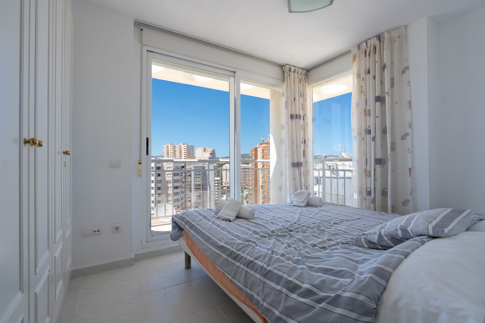 Habitación luminosa con cama doble, armario empotrado y acceso a terraza. Vistas urbanas despejadas bajo un cielo azul.