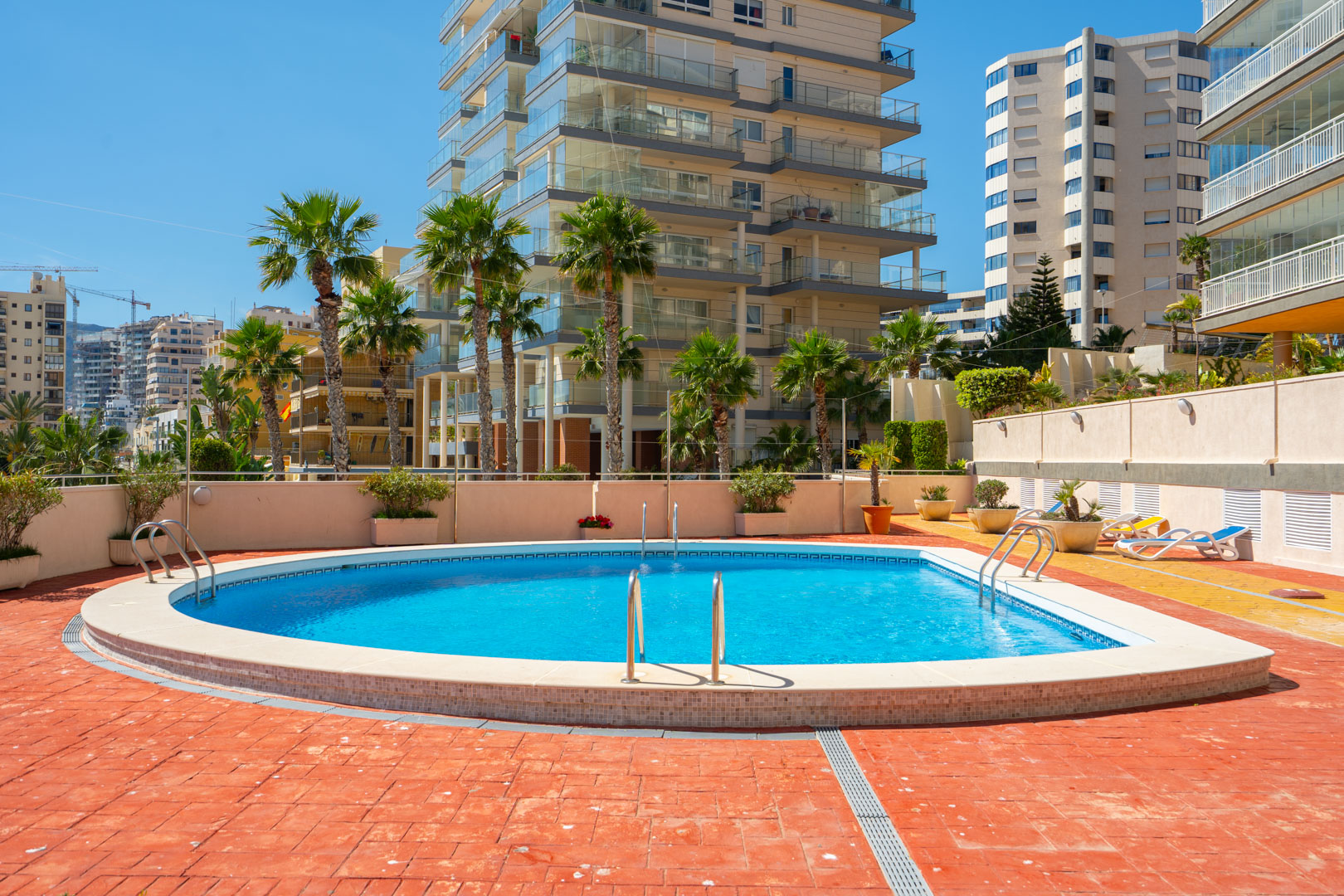 La piscina comunitaria, con sus aguas azules y escaleras de acero inoxidable, está rodeada por un solárium de baldosas de terracota y palmeras, con edificios residenciales al fondo.