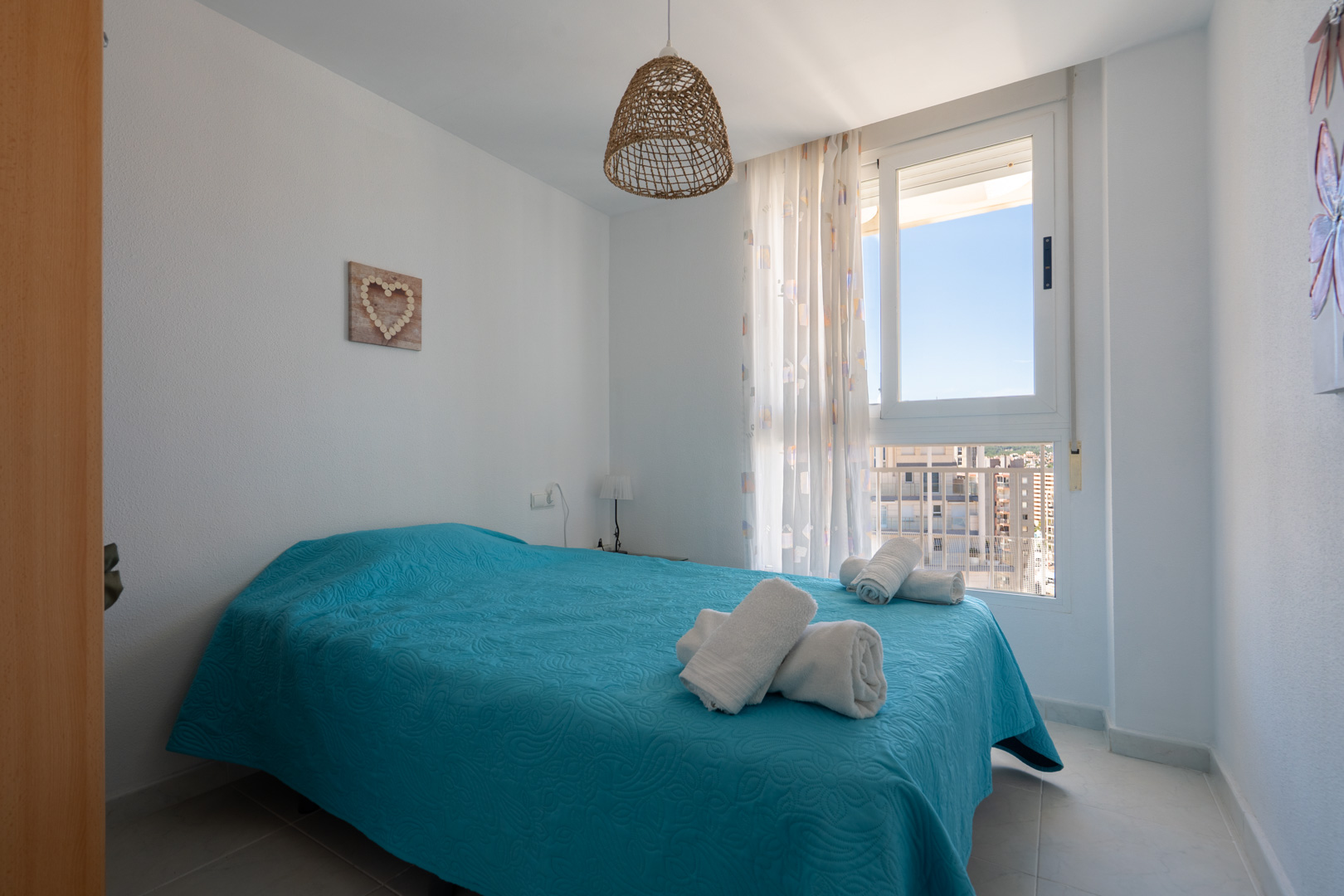 Luminoso dormitorio con cama doble vestida en turquesa, lámpara colgante de fibra natural y ventana con vistas urbanas y edificios circundantes.