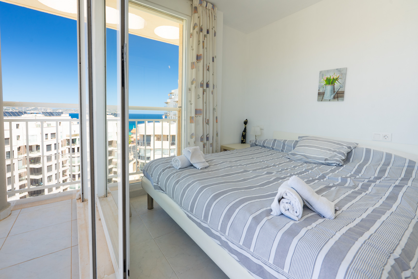 Luminoso dormitorio con cama doble y ropa de cama a rayas, mesita de noche con lámpara y acceso directo a balcón privado con vistas al mar y urbanas.