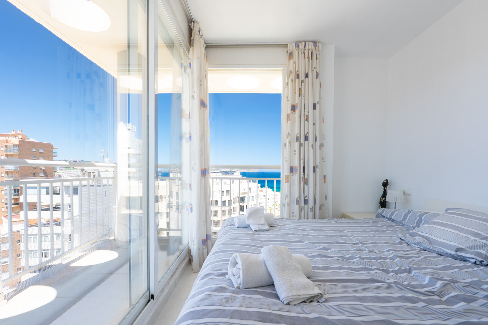 Luminoso dormitorio con cama doble y ropa de cama a rayas, ofreciendo acceso directo a una terraza con vistas panorámicas al mar y al cielo azul.