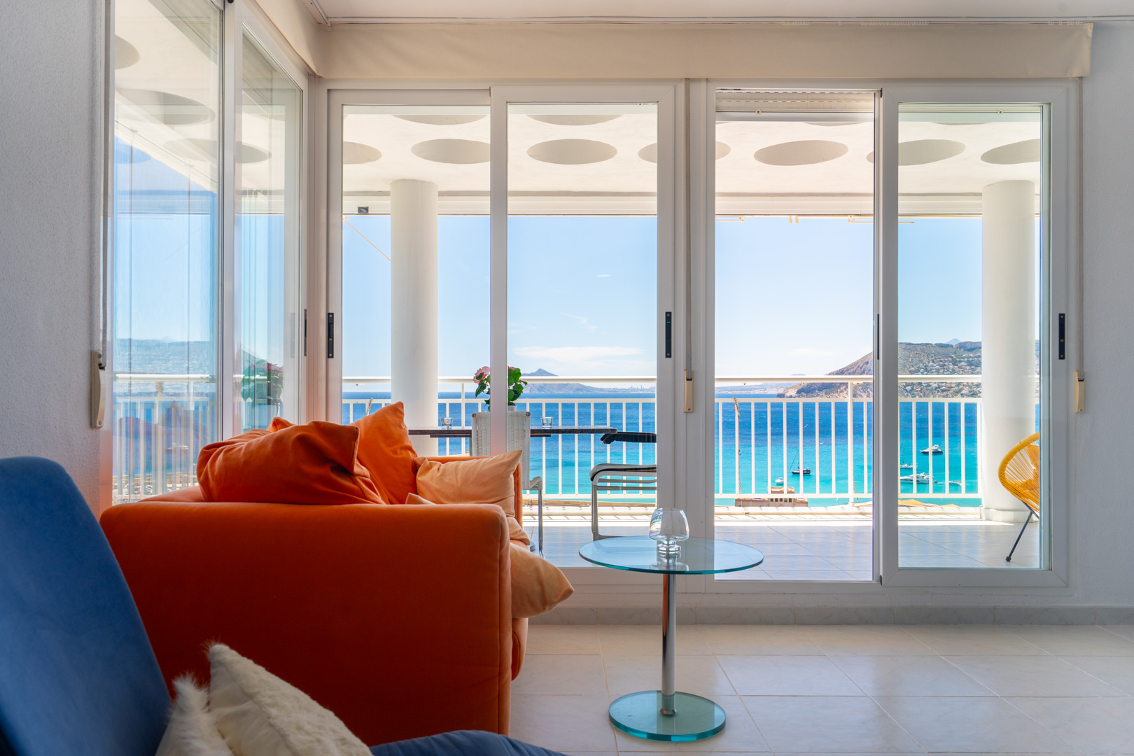 Luminoso salón con sofá naranja y mesa de cristal, con amplias puertas correderas que revelan una terraza y vistas directas al mar, barcos y montañas lejanas.