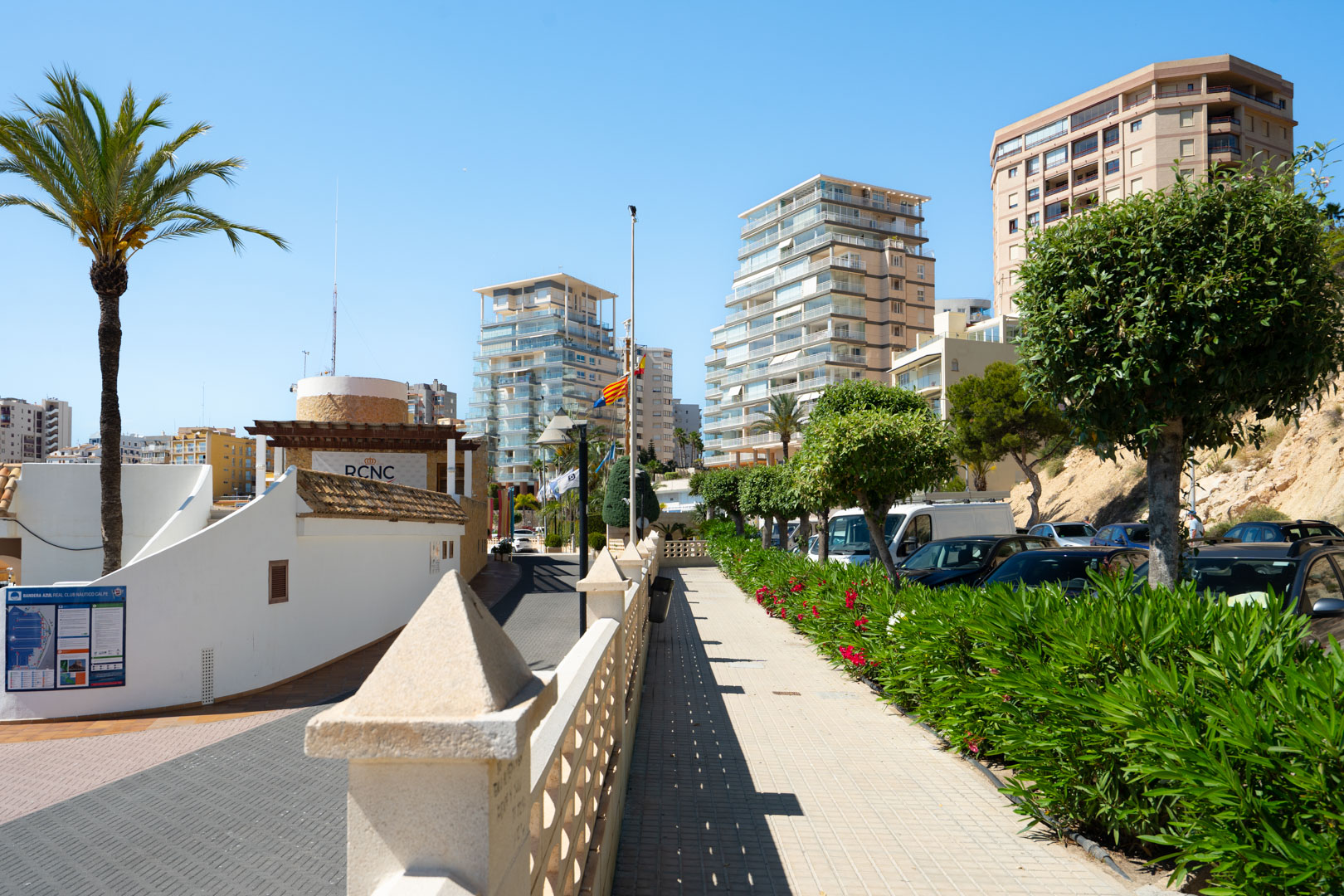 Paseo peatonal pavimentado con balaustrada de piedra y exuberantes adelfas, dirigiéndose hacia edificios modernos y la entrada del Real Club Náutico Calpe bajo un cielo azul claro.
