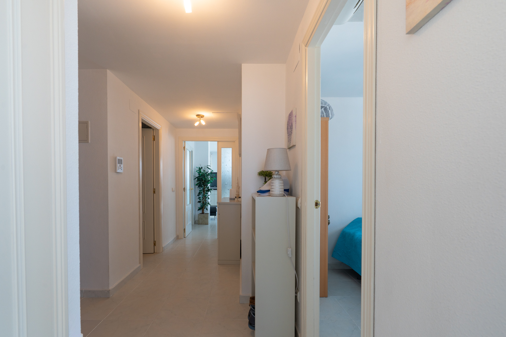 Pasillo interior con suelo de baldosas claras y paredes blancas. Varias puertas conducen a estancias, incluyendo un dormitorio visible a la derecha y una sala de estar al fondo.