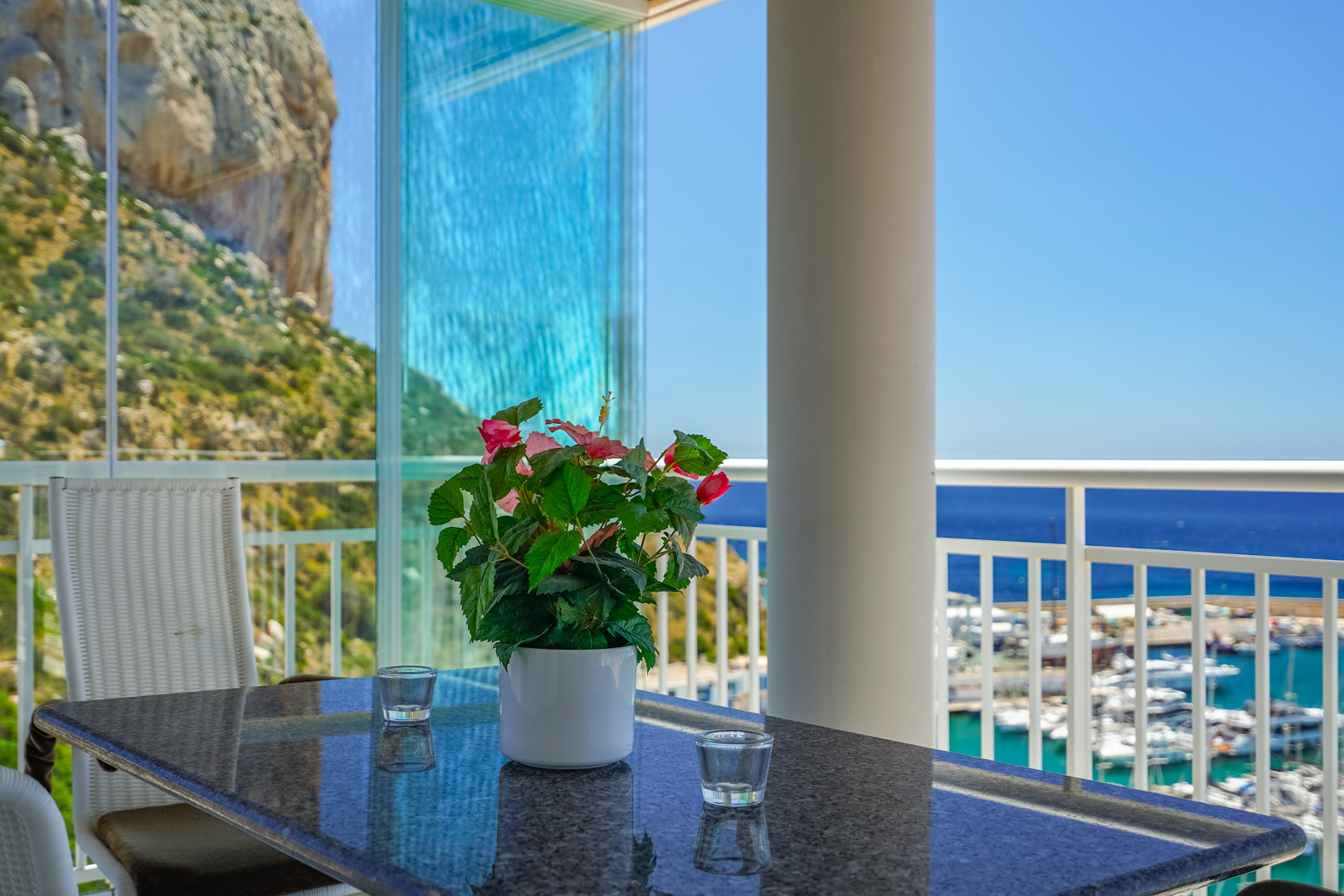 Terraza acristalada con vistas directas al Peñón de Ifach y al puerto deportivo de Calpe. Mesa de granito con planta floral y sillas de mimbre.