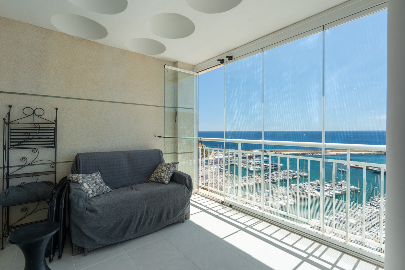 Terraza acristalada con vistas frontales al puerto deportivo y al mar, equipada con sofá y estantería de hierro forjado, bajo un techo con detalles circulares.