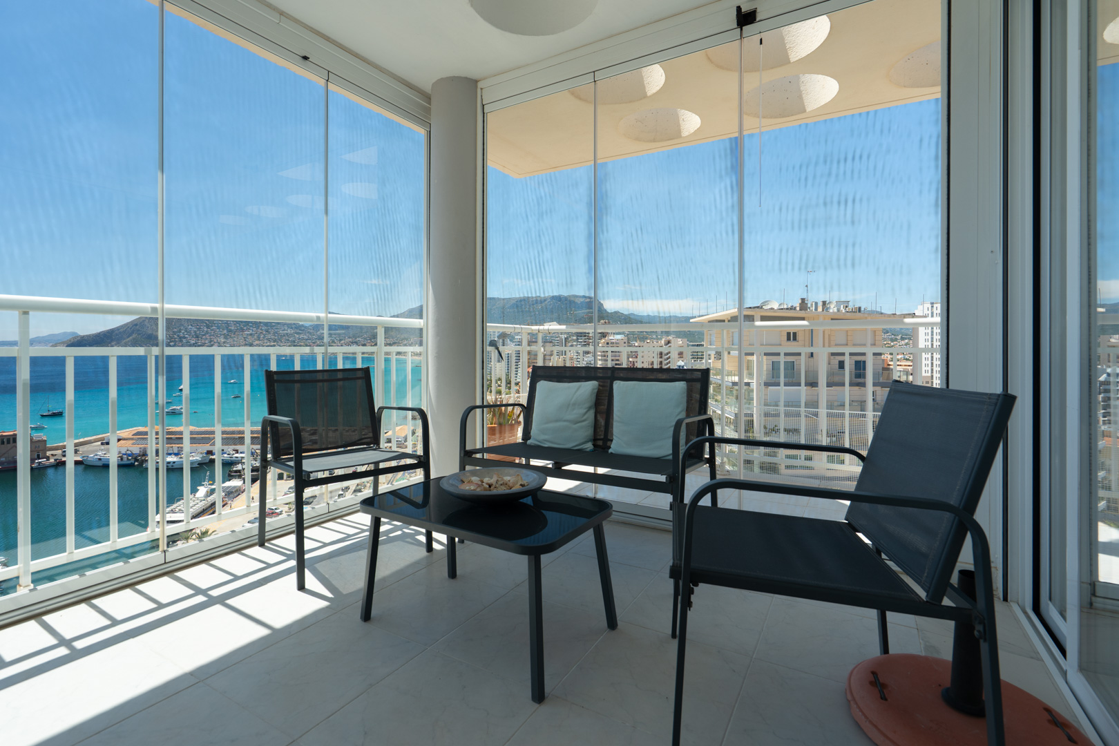 Terraza cubierta con cortinas de cristal correderas, mobiliario de exterior y vistas panorámicas al puerto deportivo de Calpe, el mar y las montañas. El suelo es de baldosas claras.