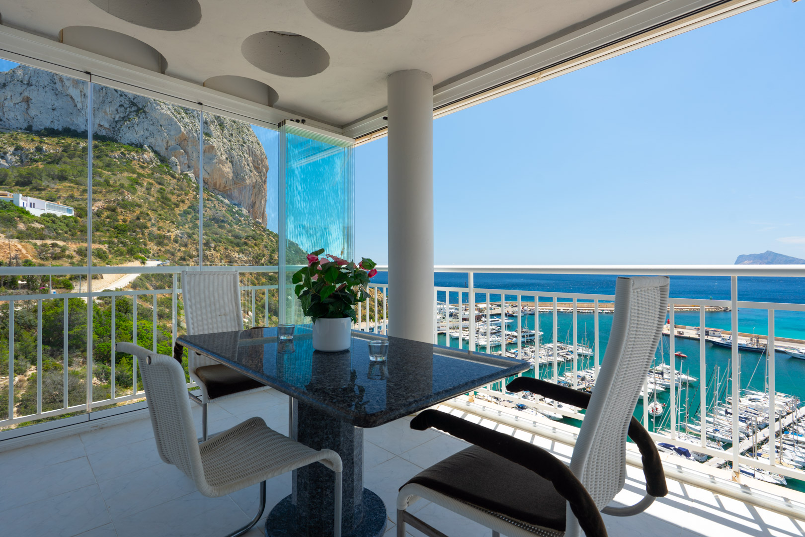 Terraza cubierta con mesa de granito y sillas modernas, equipada con cortinas de cristal, que ofrece vistas panorámicas al Peñón de Ifach y al puerto de Calpe.