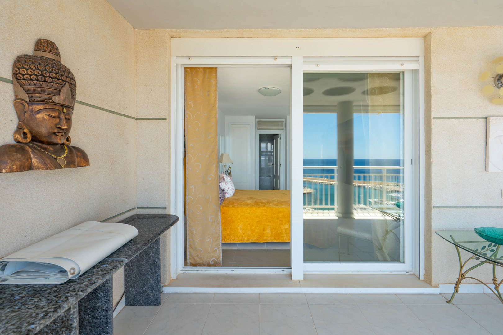 Terraza cubierta con suelo de baldosas claras y paredes texturizadas, ofreciendo acceso a un dormitorio con vistas al mar a través de puertas correderas de cristal. Incluye una consola de granito y decoración.