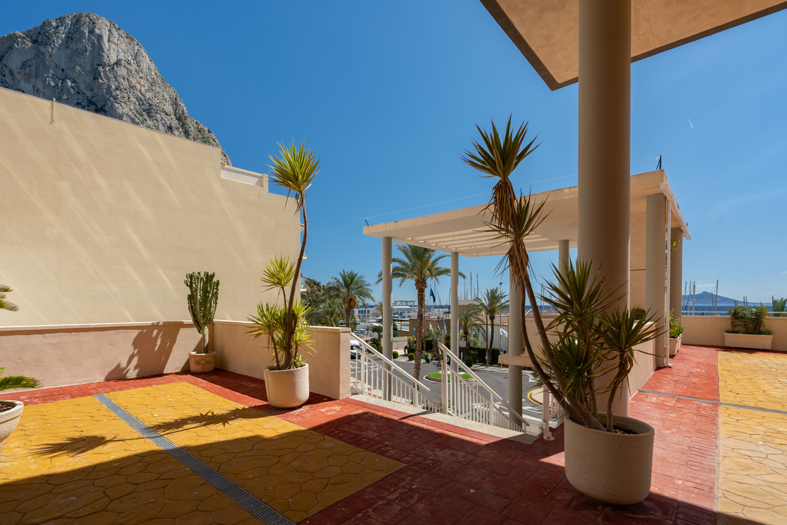 Terraza exterior con plantas mediterráneas, suelo de baldosas rojas y amarillas, y vistas al Peñón de Ifach y al puerto deportivo de Calpe.