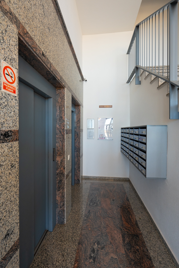 Vestíbulo del edificio con dos ascensores enmarcados en granito jaspeado, franja de suelo de granito oscuro, buzones comunitarios y escalera con barandilla metálica.
