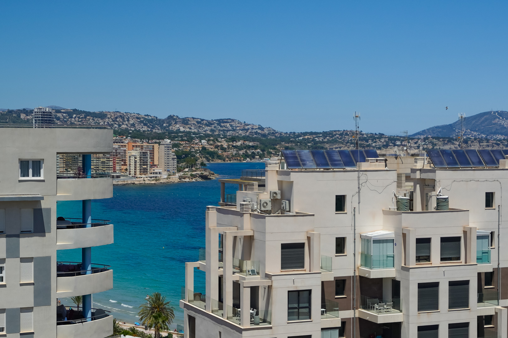 Vista desde una planta elevada de la arquitectura costera moderna, con balcones acristalados y paneles solares en los tejados, enmarcada por el mar turquesa y las colinas lejanas.