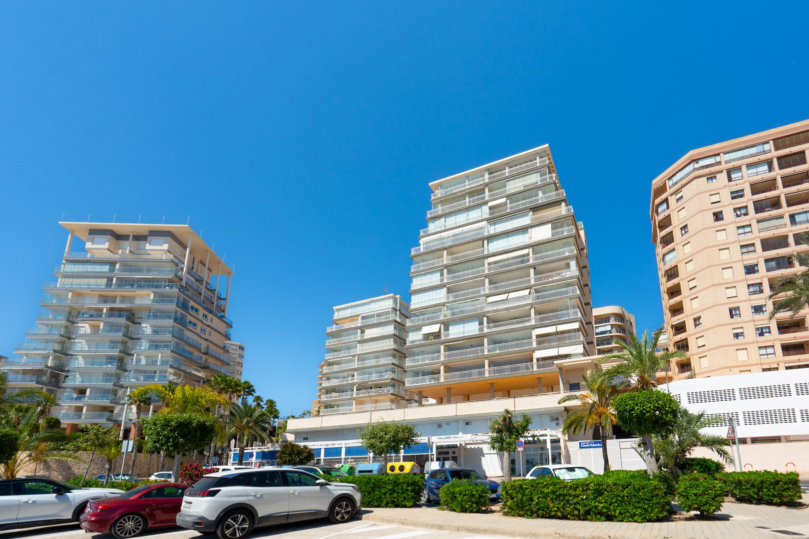 Vista exterior de modernos edificios residenciales con balcones acristalados escalonados, en un entorno urbano soleado con coches aparcados y palmeras.