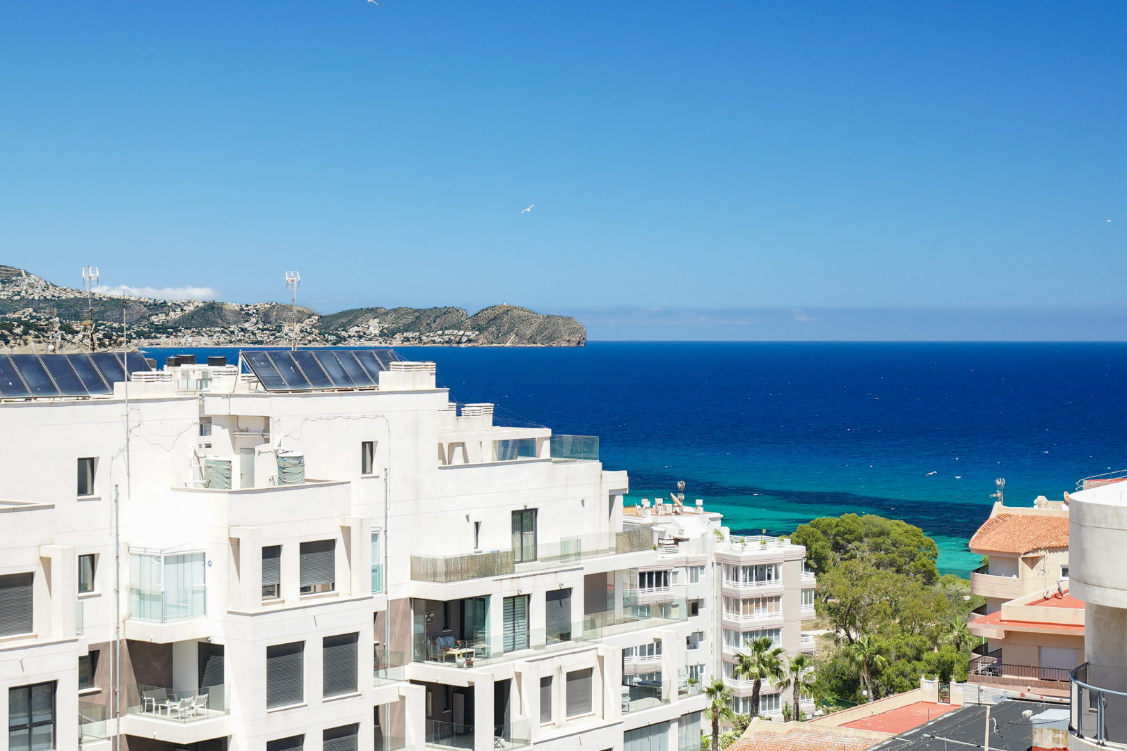 Vista panorámica de edificios residenciales blancos con balcones de cristal, paneles solares en azoteas y el azul profundo del Mediterráneo hasta la costa lejana.