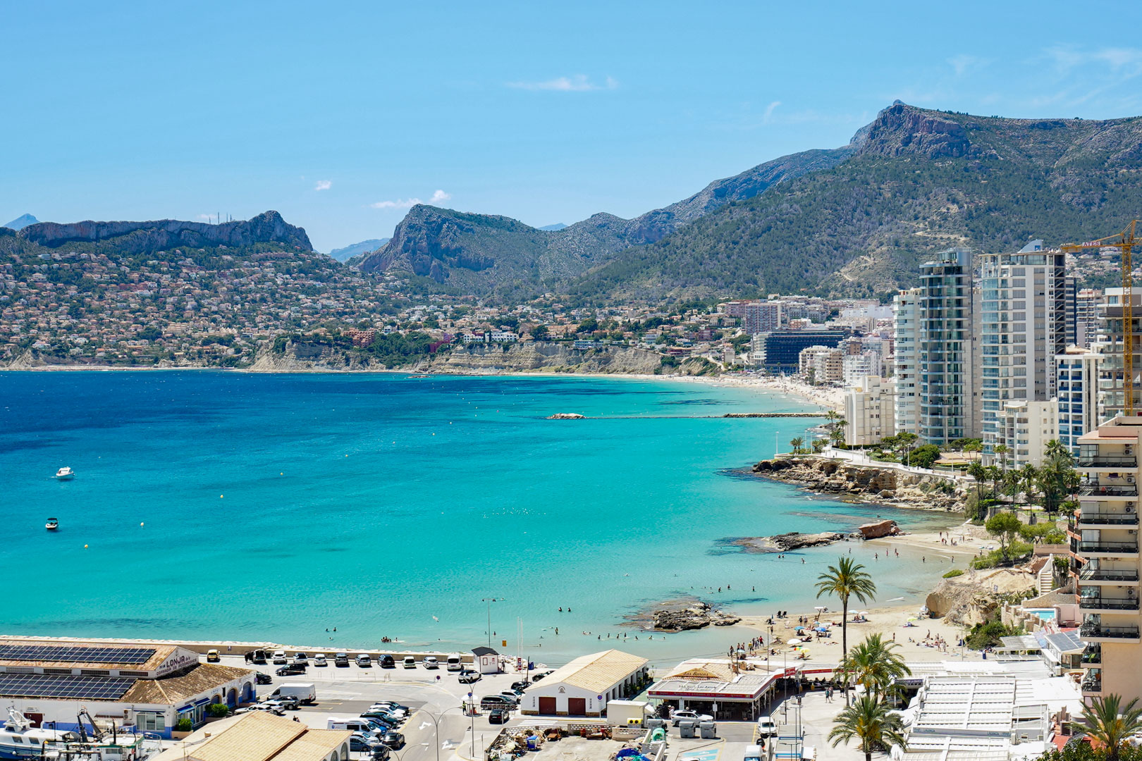 Vista panorámica de la bahía turquesa de Calpe, con playas de arena, edificios costeros y montañas lejanas bajo un cielo azul claro.