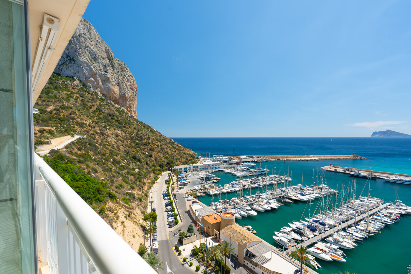Vista panorámica desde un balcón, mostrando el puerto deportivo de Calpe con numerosos barcos, el vibrante mar turquesa y el majestuoso Peñón de Ifach bajo un cielo azul claro.