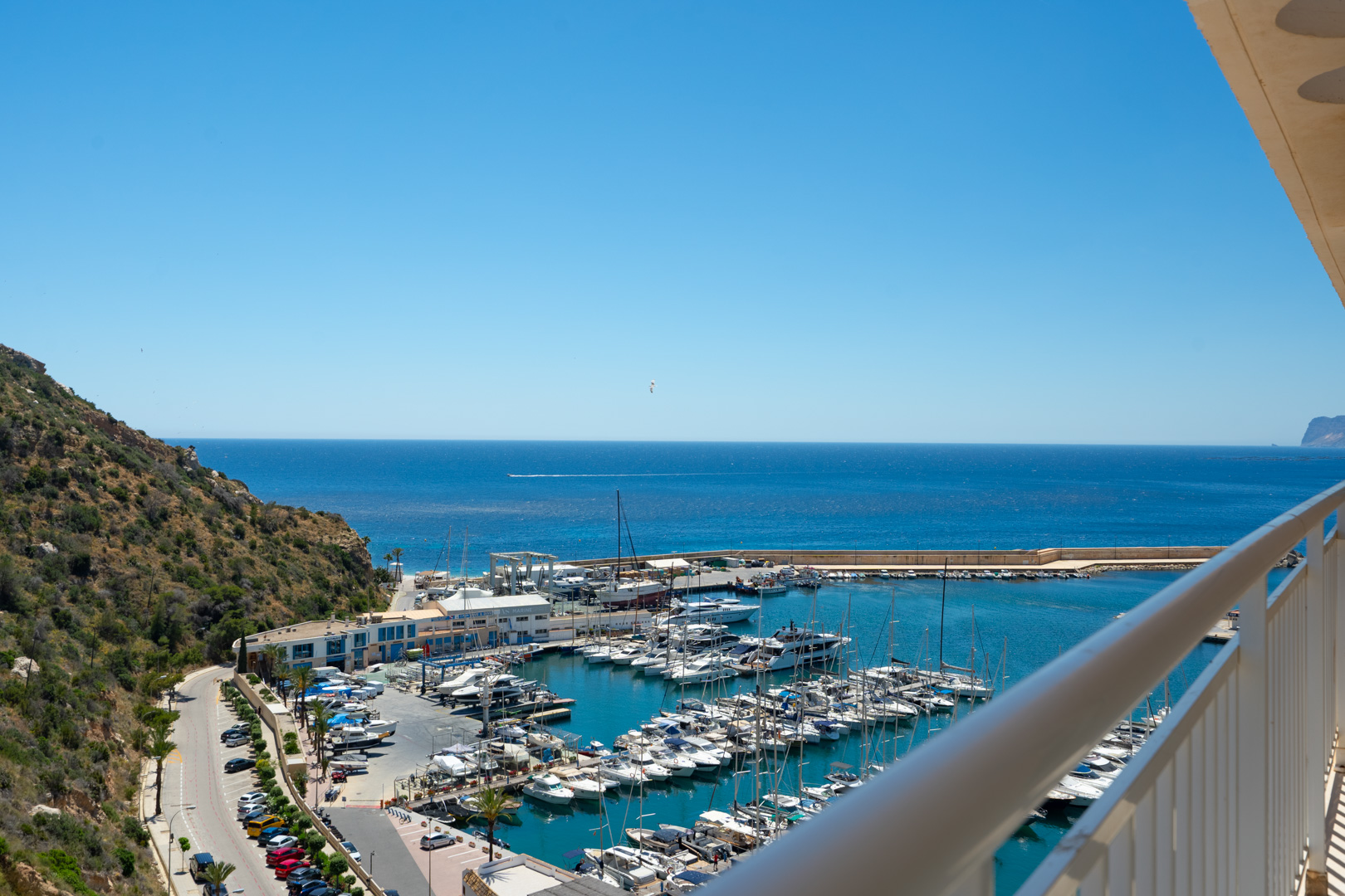 Vista panorámica desde un balcón sobre el puerto deportivo, con numerosos barcos, el mar azul profundo y una ladera verde bajo un cielo despejado.