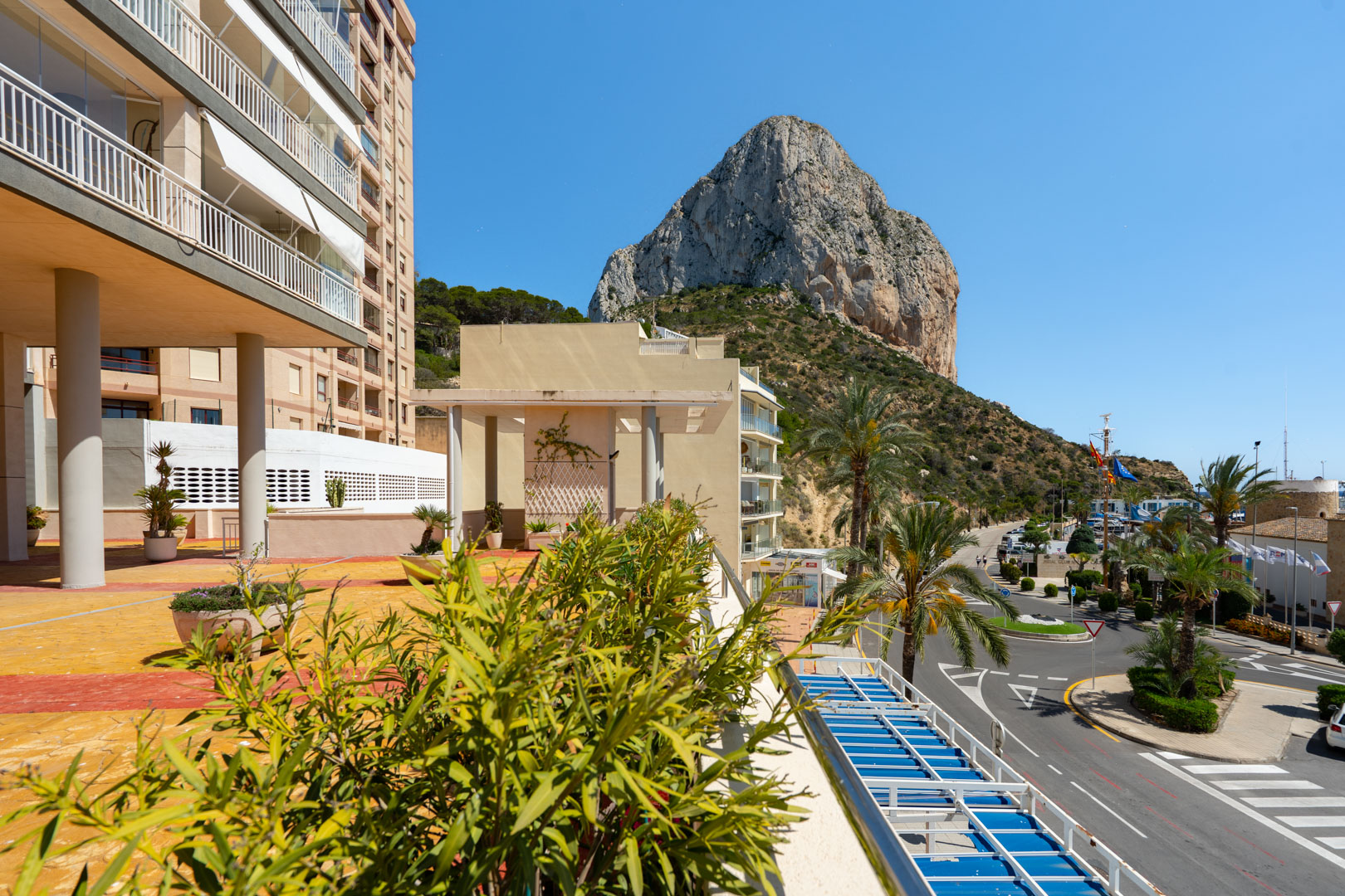 Vista panorámica desde una terraza, mostrando el icónico Peñón de Ifach, edificios circundantes y la carretera que lleva al puerto de Calpe bajo un cielo azul.