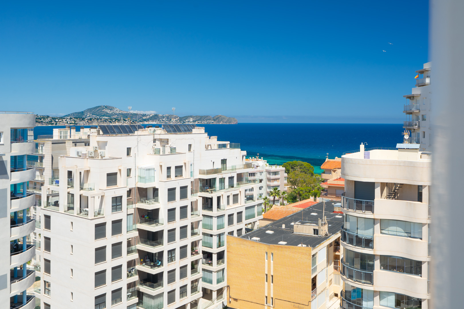 Vistas panorámicas al mar Mediterráneo y a la costa desde una planta elevada, con edificios residenciales en primer plano y montañas distantes bajo un cielo azul claro.