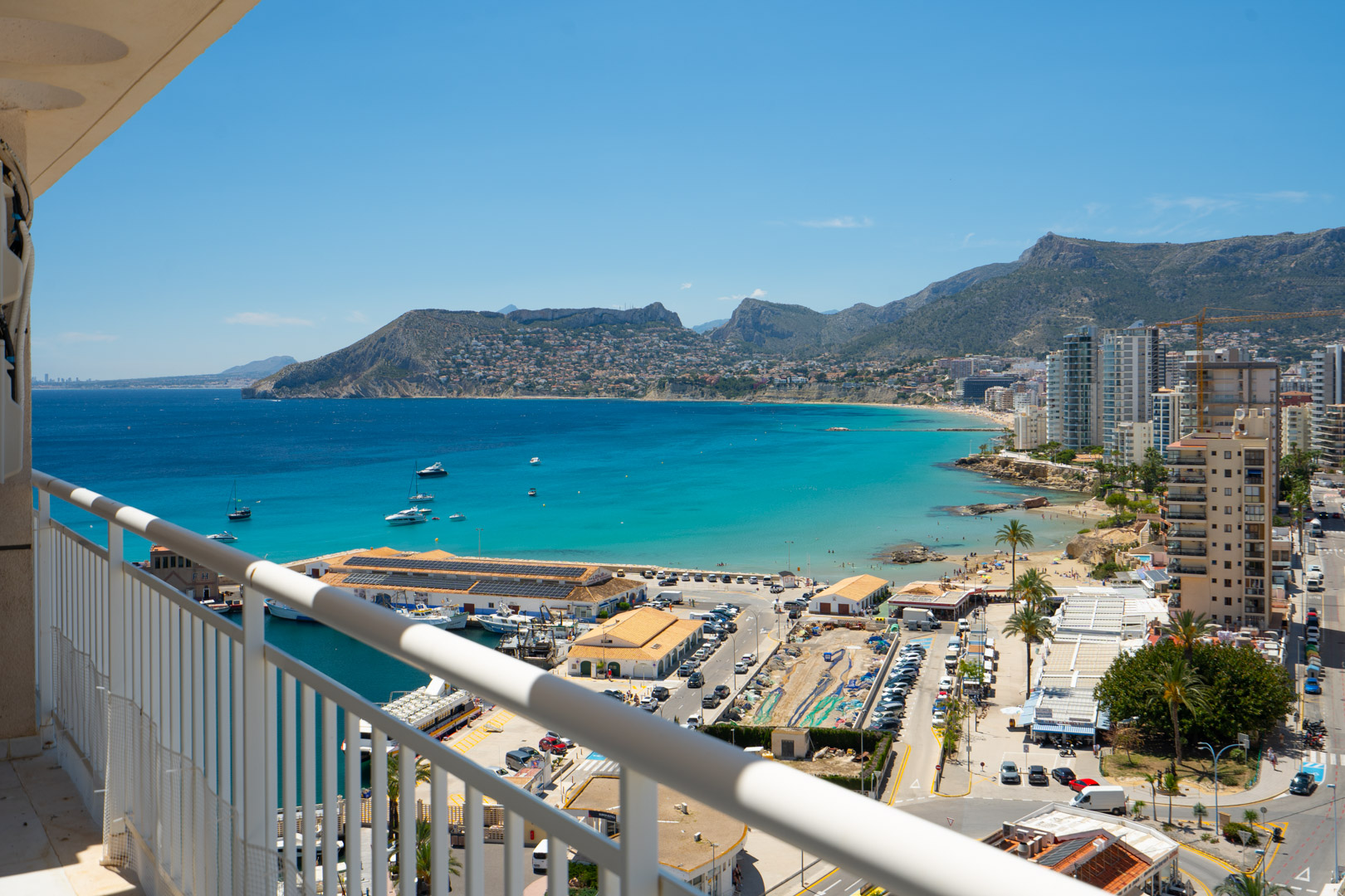 Vistas panorámicas desde la terraza al puerto de Calpe, la bahía de aguas turquesas y la costa con edificios y montañas al fondo.
