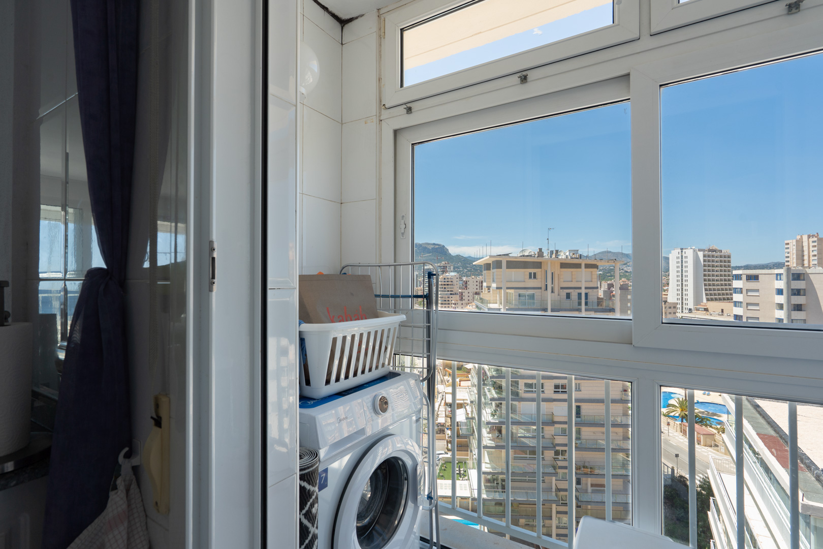 Zona de lavandería funcional con lavadora y estantería, en un espacio con paredes alicatadas y amplios ventanales que enmarcan vistas urbanas a edificios, montañas y una piscina.