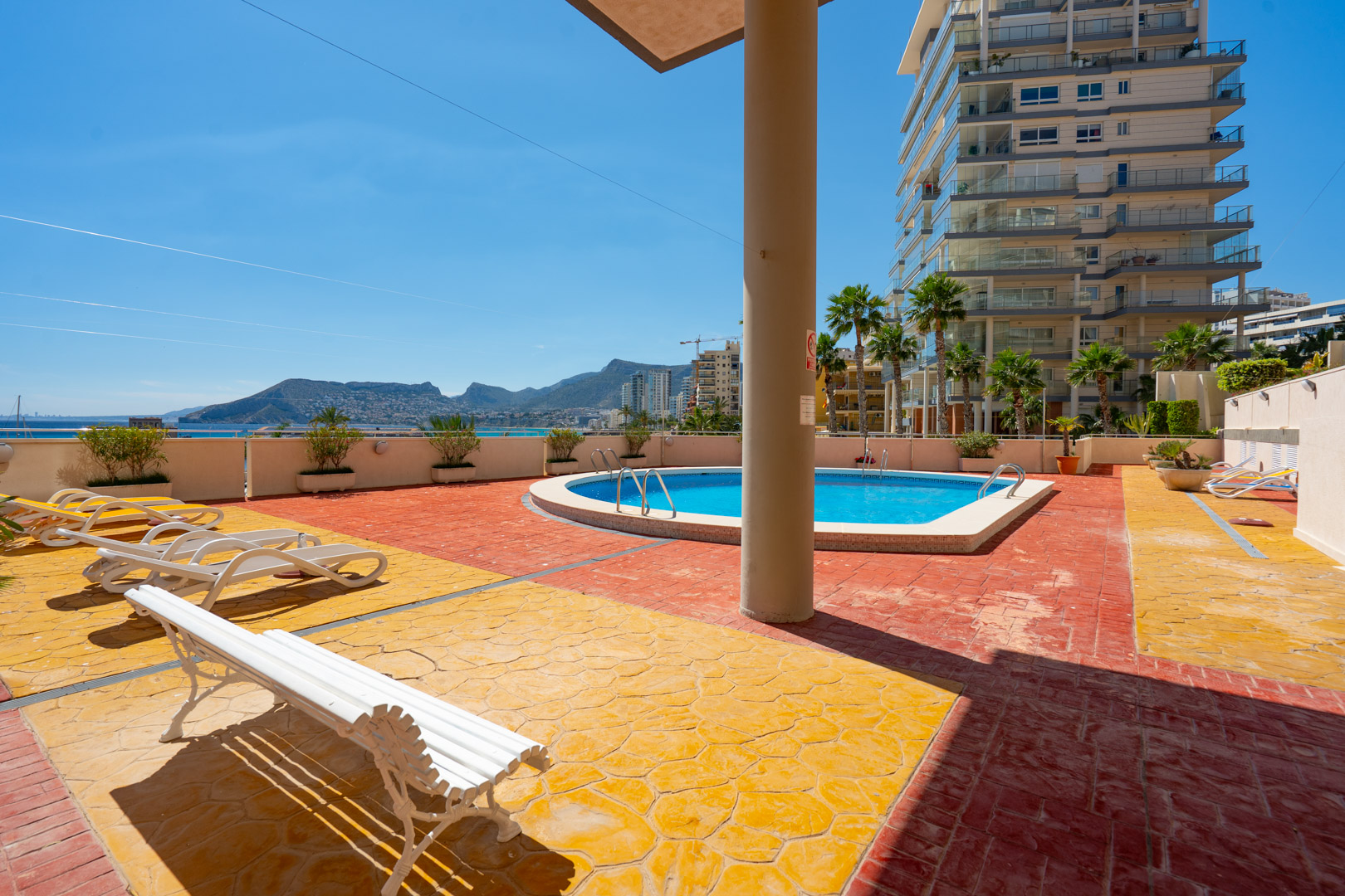 Zona de piscina comunitaria con pavimento texturizado amarillo y terracota, tumbonas y un banco blanco. Ofrece vistas al mar, montañas y al moderno edificio residencial con palmeras.