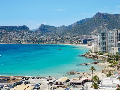 Amplia panorámica costera de la bahía de Calpe, con aguas turquesas, playas de arena, edificios modernos y montañas lejanas. El puerto con paneles solares se aprecia en primer plano.