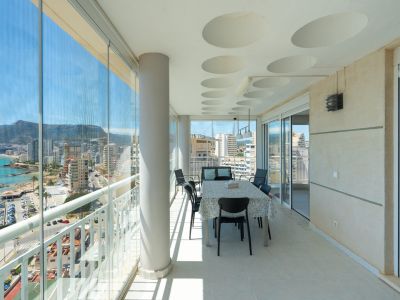 Amplia terraza acristalada con comedor y vistas panorámicas al mar, puerto y montañas de Calpe. Destacan las cortinas de cristal, el techo con detalles circulares y el suelo de gres.