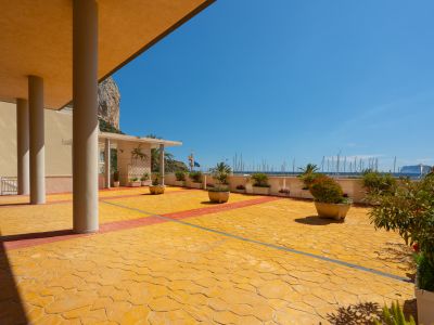 Amplia terraza comunitaria con pavimento amarillo texturizado y detalles rojos, área cubierta con robustas columnas y vistas panorámicas a la marina y al Peñón de Ifach.