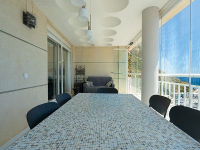 Amplia terraza cubierta con mesa de comedor de mosaico, asientos y cortinas de cristal con vistas panorámicas al puerto, el mar y las montañas.