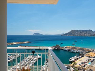 Amplias vistas al mar y al puerto deportivo desde el balcón, con numerosos barcos, la bahía turquesa y montañas lejanas bajo un cielo azul claro.