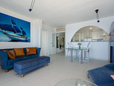 Amplio salón-comedor con sofá azul, mesa de cristal y sillas transparentes, complementado por un gran cuadro de velero. Una arcada conecta con la moderna cocina semiabierta.