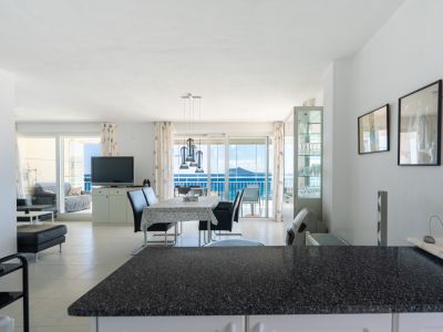 Amplio salón-comedor de planta abierta con lámpara colgante moderna sobre la mesa, suelo de baldosas claras y acceso directo a una terraza con vistas panorámicas al mar a través de puertas correderas de cristal.