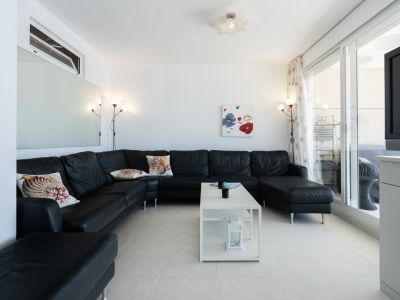 Amplio salón con un gran sofá seccional negro y cojines decorativos, suelo de baldosas blancas y mesa de centro moderna. Las puertas correderas de cristal dan a una terraza.