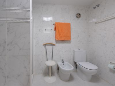Baño con azulejos efecto mármol blanco, equipado con inodoro, bidé y una práctica zona de ducha con barra de apoyo. Incluye un reloj de pared y perchas para toallas.