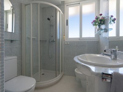 Baño con ducha de esquina, inodoro, bidé y lavabo con encimera de mármol. Paredes revestidas con mosaico azul claro y ventanas que aportan luz natural.