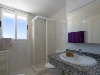 Baño funcional con azulejos efecto mármol blanco, encimera de granito gris y lavabo ovalado. Incluye cabina de ducha de esquina y ventana con luz natural.