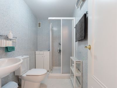 Baño funcional con paredes de mosaico azul claro, equipado con inodoro, lavabo de pedestal y cabina de ducha. Incluye un pequeño mueble de almacenamiento y estanterías auxiliares.