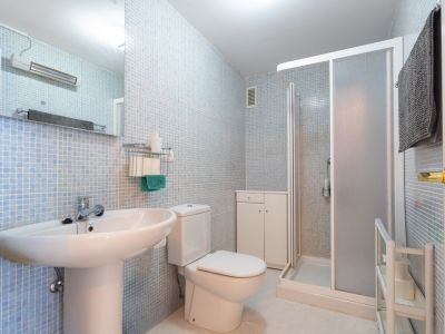 Baño funcional con paredes de mosaico azul claro, lavabo de pedestal, inodoro, ducha con mampara de cristal esmerilado y mueble auxiliar blanco para almacenamiento.