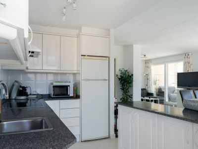 Cocina equipada con armarios blancos panelados, encimeras de granito oscuro moteado y fregadero de acero inoxidable, abierta al salón mediante una barra americana y con luz natural de la terraza.