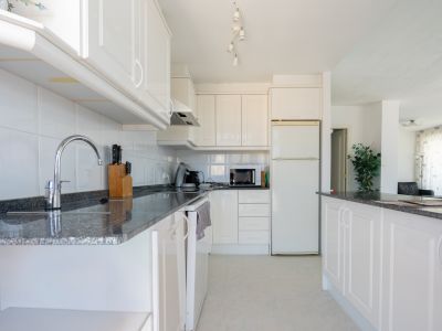 Cocina funcional con mobiliario blanco, encimeras de granito oscuro y azulejos blancos. Equipada con electrodomésticos esenciales y barra americana que conecta con el salón.